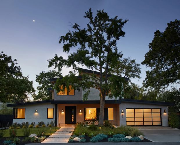 Twilight exterior view of a contemporary two-storey residence with a low-pitched roof and broad single-storey wings. The home has smooth light-grey rendered walls, warm illuminated windows, a dark-framed entry, and a large gridded garage door. A landscaped front garden with ornamental grasses, low shrubs, boulders, and a paved path leads to the entrance, while mature trees partially screen the facade.