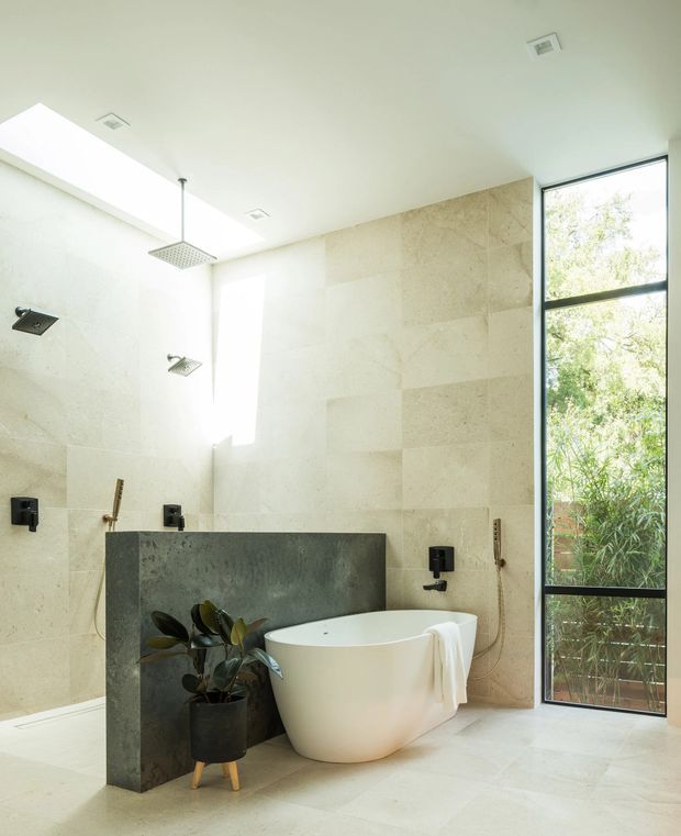 Bright, spacious contemporary bathroom with a freestanding white bathtub, large-format pale stone wall and floor tiles, dark stone privacy screen, multiple black wall-mounted fixtures, a rainfall shower, and a tall black-framed window overlooking greenery. A potted plant adds a natural accent.