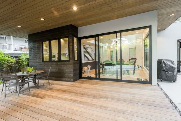 A covered timber deck or terrace adjoining a modern home, with wide timber decking, an outdoor dining setting, and a large black-framed glazed opening into a bright interior. The extension combines dark horizontal timber cladding, white rendered walls, timber soffit lining, and a visible internal stairway and living area. A covered barbecue or appliance sits at the right edge.