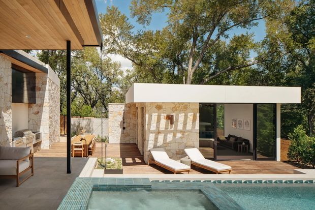 A contemporary single-storey pool courtyard framed by mature trees. The scene features pale natural-stone walls, a crisp white flat-roofed pavilion with extensive dark-framed glazing, warm timber soffits and decking, and a tiled swimming pool in the foreground. Outdoor loungers, a dining setting, and a built-in barbecue create a refined indoor-outdoor entertaining area.