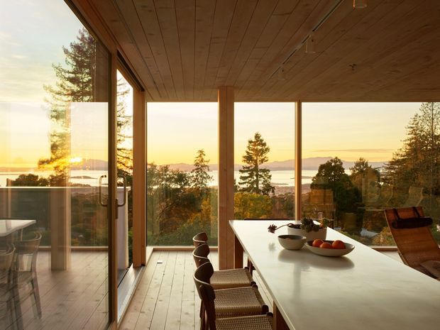 A warm, light-filled dining area with a long white dining table and woven chairs, enclosed by expansive floor-to-ceiling glazing. Timber-framed glass doors and windows open views toward a wooded hillside and distant water at sunset. The ceiling and floor use exposed timber, creating a calm, nature-connected interior.