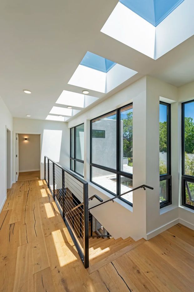 Bright contemporary stair landing and hallway with wide-plank timber flooring, white walls, black metal-and-cable stair balustrade, large black-framed windows, and a row of rectangular skylights bringing in abundant daylight. The stair descends beside the glazing, with views to greenery outside.