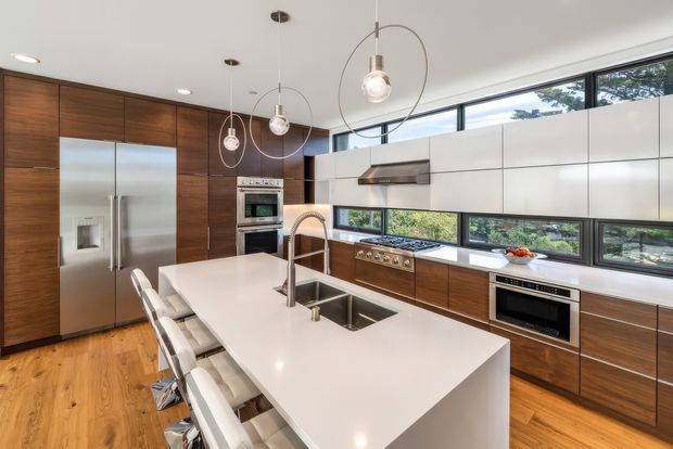 Bright contemporary kitchen with a large white island, integrated stainless-steel sink and tap, warm timber cabinetry, stainless-steel appliances, and timber flooring. Linear clerestory-style windows provide abundant natural light, while globe pendant lights hang above the island.