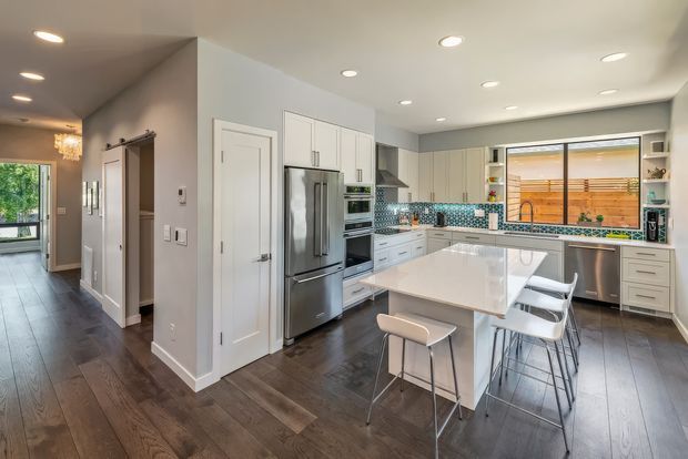 Bright, spacious contemporary kitchen with white full-height cabinetry, stainless-steel appliances, a large white island with integrated seating, and a blue-grey mosaic tile splashback. Dark timber flooring continues into the adjacent hallway, which includes recessed ceiling lighting and a decorative pendant. A large rear window overlooks a timber privacy screen and outdoor area.