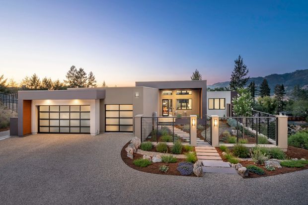 A modern single-storey residence photographed at dusk, featuring a broad flat-roofed facade, two integrated garage doors, warm grey rendered walls, dark-framed glazing, and a raised entry reached by a stepped landscaped path. Metal mesh fencing and low-maintenance planting define the front garden, with wooded hills visible in the background.