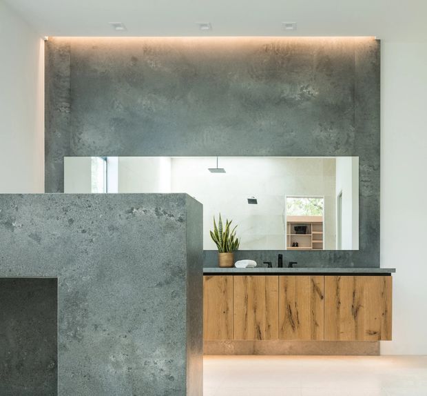 Contemporary bathroom vanity area featuring a large frameless mirror, a floating natural timber vanity with dark fixtures, and a dramatic textured grey concrete or stone feature wall and partition. Concealed warm LED lighting adds a refined, softly illuminated effect, while a potted plant provides a small natural accent.