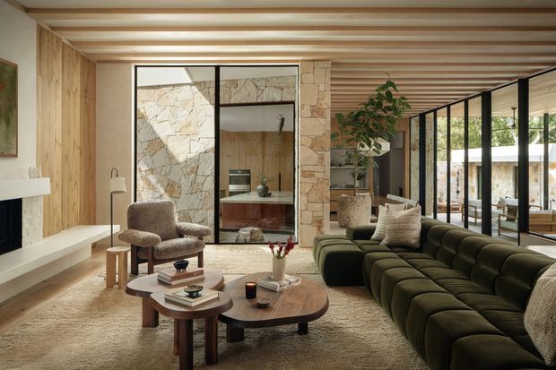 Warm, refined living room with a large deep olive-green sectional sofa arranged around layered rounded timber coffee tables. The space features exposed light timber wall and ceiling boards, a horizontal slatted timber ceiling, natural irregular stone wall surfaces, black-framed internal glazing, a built-in white fireplace shelf, and views into an adjacent courtyard-like area. Neutral upholstered lounge chairs, a textured rug, indoor planting, and understated decor complete the inviting interior.