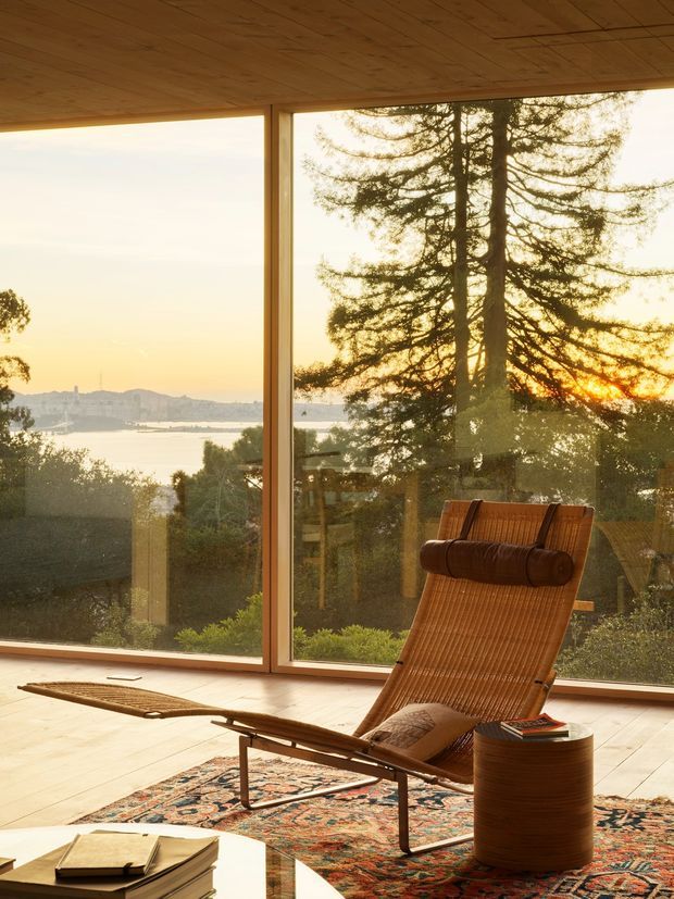 A warm, serene interior lounge or sunroom with expansive floor-to-ceiling glazing overlooking a waterfront or bay at sunset. The room features a timber-lined ceiling, pale timber flooring, a vintage woven reclining lounge chair, patterned area rug, and a small cylindrical side table. Reflections in the glazing reveal additional interior furnishings, while the surrounding landscape and water form the exterior view.