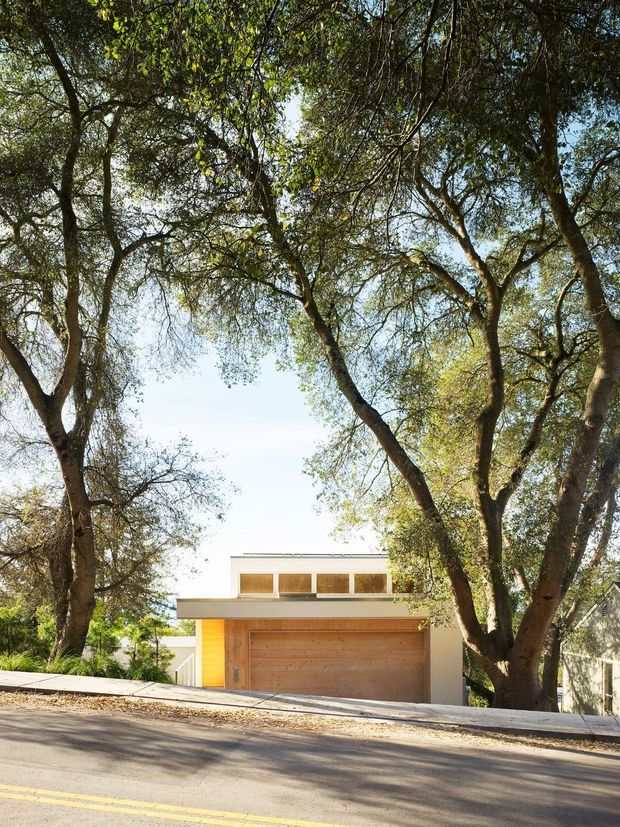 Street-level exterior view of a compact contemporary residential garage or carport structure set beneath mature trees. The building has a broad timber garage door, warm timber-clad side wall, pale rendered walls, and a simple flat roof with a row of upper windows. A sloping paved street and sidewalk occupy the foreground, with dense leafy landscaping framing the facade.