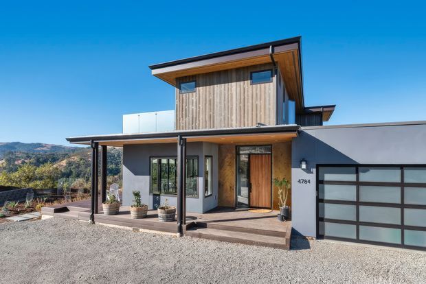 Exterior view of a contemporary two-storey residence on a sloping site, featuring vertical timber cladding, dark painted walls, broad roof overhangs, a covered entry porch, large glazed openings, and an integrated double garage. Gravel landscaping and distant hills are visible.