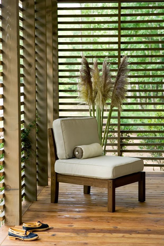 A calm, light-filled covered outdoor sitting area with a cream upholstered lounge chair on a timber deck. Vertical timber posts and horizontal timber slatted screening create a partially enclosed space, with greenery visible beyond. A vase of tall dried grasses adds a natural decorative element, and sandals rest on the deck.