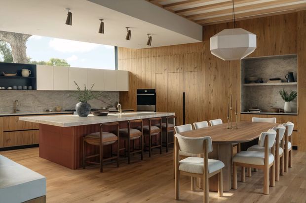 Warm, open-plan kitchen and dining area with extensive timber wall panelling and wood flooring. The kitchen features a large island with dark wood cabinetry, a pale stone benchtop and integrated appliances, while a long timber dining table is surrounded by upholstered dining chairs. Open shelving, a marble-look splashback, contemporary pendant lighting and broad glazing complete the refined interior.