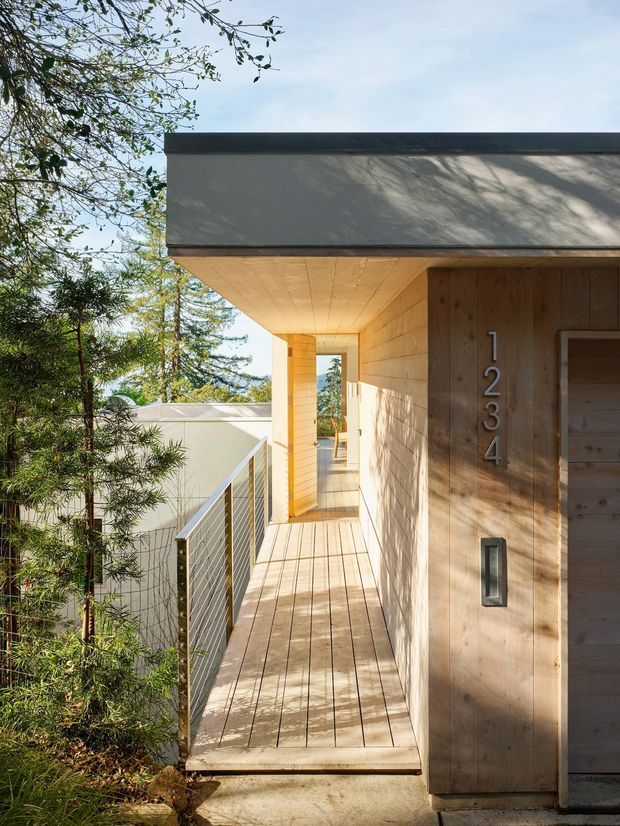 A sunlit covered exterior walkway or balcony runs alongside a contemporary timber-clad building. The passage has a pale timber plank floor and ceiling, warm vertical timber wall cladding, a slim dark roof edge, and a metal cable-style balustrade. Large numerals mark the entrance beside a narrow vertical light fitting. Trees and another light-coloured structure are visible beyond the railing.