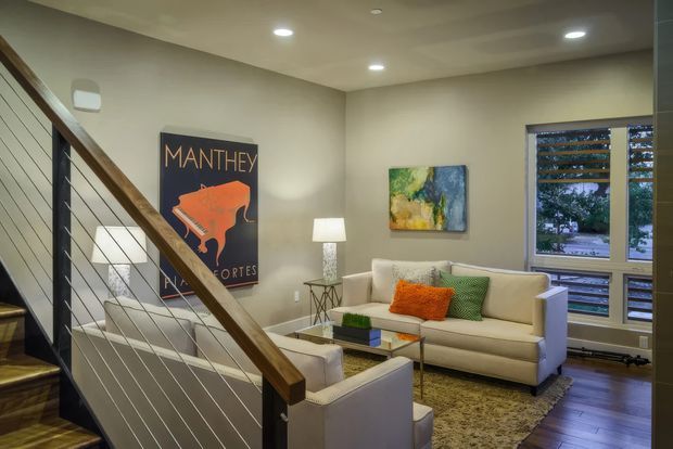 A warm, contemporary living room viewed from a staircase. The room features a cream sofa and armchairs, a glass-topped coffee table, a floor lamp, wall art, recessed ceiling lights, and a large window with horizontal blinds. The staircase has dark timber treads, a timber handrail, and slim horizontal metal balusters. Dark timber flooring and a patterned area rug add contrast to the neutral walls and upholstery.