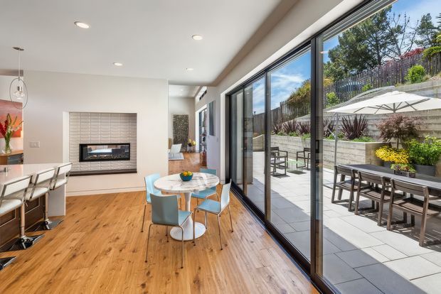 Bright, open-plan contemporary living and dining area with warm timber flooring, a compact dining table and blue upholstered chairs, and a linear kitchen bench at left. A tiled feature wall surrounds a built-in fireplace, while extensive black-framed sliding glass doors open to a paved outdoor terrace with dining furniture, planting, and umbrellas. The space has recessed ceiling lighting and strong indoor-outdoor connection.