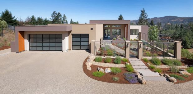 Single-storey contemporary residence viewed from the front, with a flat-roofed geometric form, warm beige rendered walls, dark-framed glazed garage doors, and a raised entry terrace reached by a central concrete stepping-stone path. Low drought-tolerant planting, gravel and boulders form the landscaped foreground, with wooded hills visible beyond.