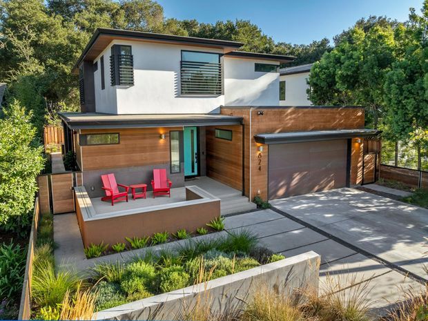 A contemporary two-storey residence viewed from the street, with crisp white rendered upper walls, horizontal dark-framed windows, warm timber-clad lower walls, and a low-pitched flat roofline. The home includes an integrated double garage, a paved driveway, and a recessed front terrace with bright red outdoor chairs and a small table. Layered planting and concrete garden walls frame the property.