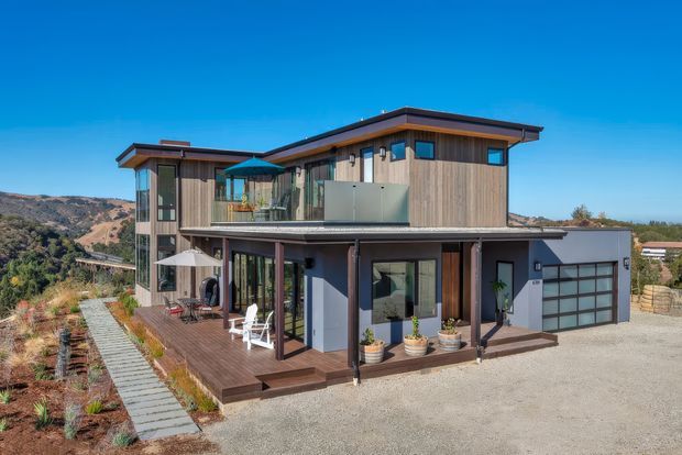 Exterior view of a contemporary multi-level residence on a sloped site, featuring vertical timber cladding, dark metal roof planes, extensive glazing, a glass balcony balustrade, and a broad timber deck with outdoor seating. A garage is integrated at ground level, with landscaped planting and a gravel approach surrounding the home.