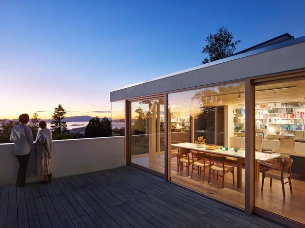 A modern flat-roof residence viewed from a spacious rooftop or elevated timber deck at dusk. Full-height glazed sliding doors reveal a warm, open dining and kitchen area with a long wooden dining table, chairs, pendant lighting, shelving, and cabinetry. A low parapet frames broad water and landscape views, while two people stand at the deck edge. The architecture features clean horizontal lines, dark timber decking, timber-framed glazing, and a restrained neutral facade.