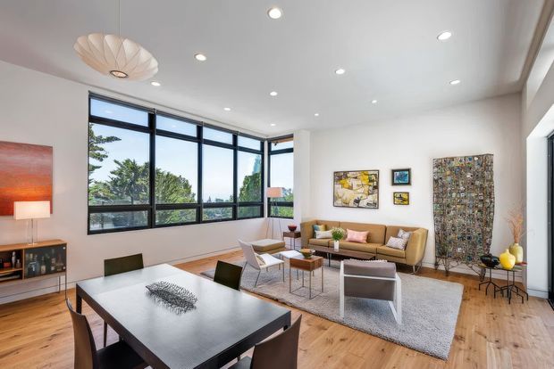 A bright, open-plan contemporary living and dining area with expansive black-framed corner windows, pale timber flooring, white walls, and recessed ceiling lights. A dark rectangular dining table occupies the foreground, while a tan sofa, light armchairs, coffee table, large neutral rug, artwork, and decorative lighting create a refined lounge zone. The room has generous glazing and views to greenery outside.
