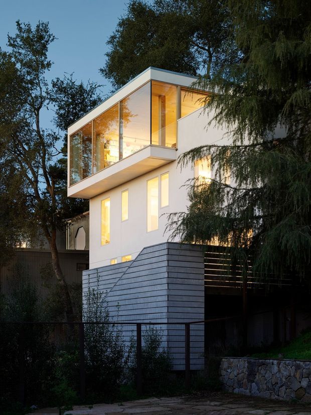 Photograph of a contemporary multi-storey house viewed from outdoors at dusk, set among mature trees. The building has crisp white rendered walls, extensive floor-to-ceiling glazing at the upper level, warm timber-lined soffits and interiors visible through the windows, projecting box-like forms, and a dark horizontal slatted privacy screen over a lower area. A stone retaining wall and landscaped garden sit in the foreground.
