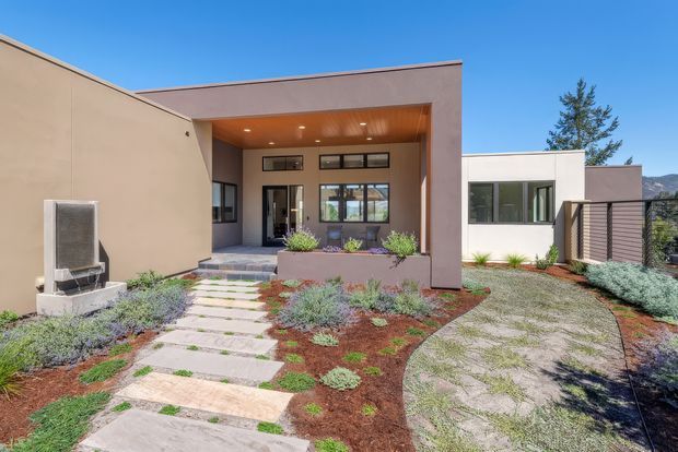 Contemporary single-storey home exterior with a minimalist geometric composition, flat rooflines, smooth rendered walls, and a recessed entry porch with a timber-lined ceiling. The front garden features drought-tolerant planting, gravel, and irregular concrete stepping stones leading to the entrance. Large dark-framed windows and a slatted privacy screen add contrast, with distant hills visible beyond the property.