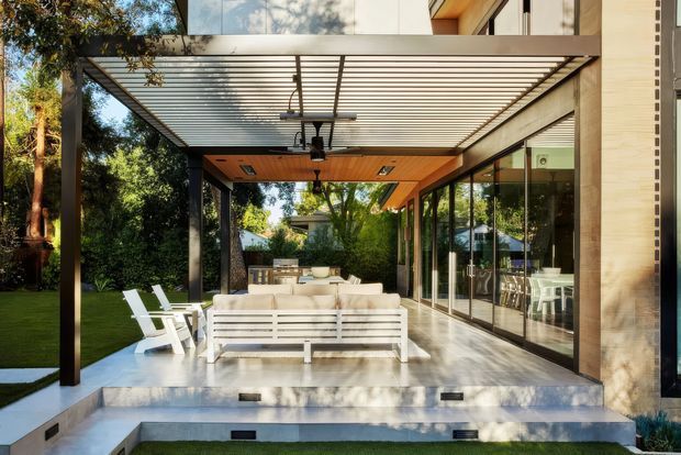 A sophisticated covered outdoor terrace adjoining a modern glass-walled residence. The space features a white outdoor lounge arrangement, dining area, timber-lined ceiling, dark structural posts, a horizontal louvred pergola roof, and broad concrete steps leading toward a manicured garden. The glazing reveals an interior dining area, while mature trees create a leafy suburban setting.
