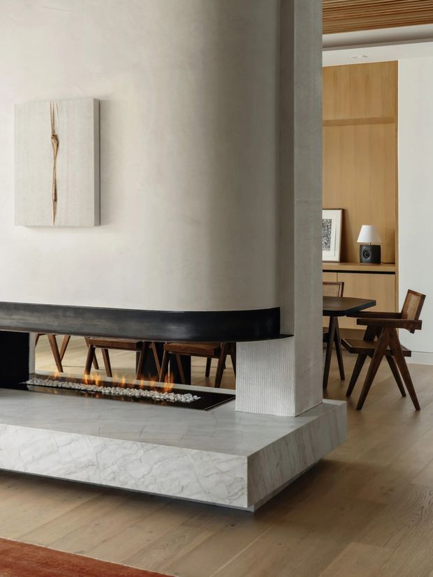 Refined open-plan interior featuring a sculptural marble-clad fireplace hearth with a long black firebox, a textured pale plaster partition, timber flooring, and a dining area with mid-century wooden chairs visible beyond. Warm timber wall panelling and a slatted timber ceiling add natural detail, while minimalist artwork and a small table lamp complete the restrained scheme.