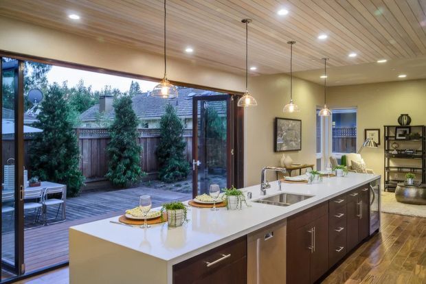 A bright, open-plan kitchen with a large white island, integrated sink and dishwasher, dark wood cabinetry, and multiple glass pendant lights. Full-height glazed doors open onto a timber deck and landscaped courtyard, while a dining and living area with shelving is visible beyond. The ceiling has warm horizontal timber slats and the floor appears to be timber flooring.