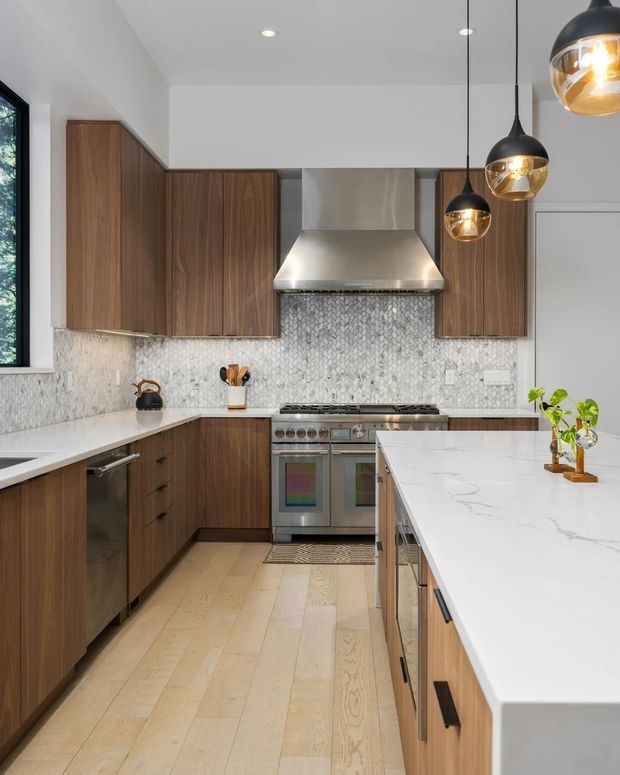 A contemporary galley-style kitchen with warm walnut cabinetry, a large white stone island, pale timber flooring, and a light grey mosaic tile splashback. Stainless steel appliances, a sculptural rangehood, and three black-and-brass pendant lights create a refined focal area. The kitchen is bright, compact to medium-sized, and efficiently arranged.