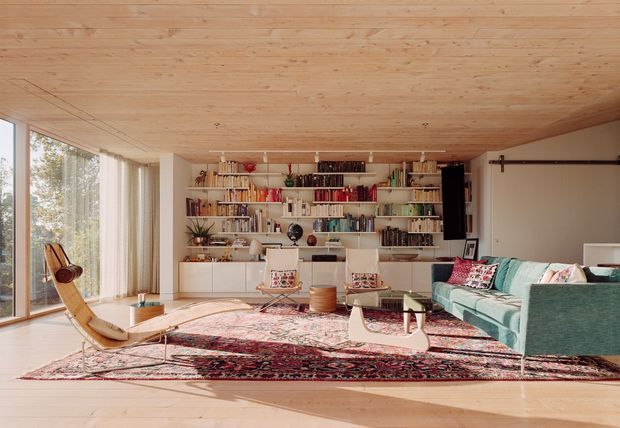 A warm, light-filled living room with a low timber-lined ceiling and matching timber floor. Full-height glazing with sheer curtains occupies the left side, while a large built-in white bookcase and cabinetry spans the rear wall. The room includes a turquoise sectional sofa, patterned rugs, a timber lounge chair, a low coffee table, and eclectic books and decorative objects. A wall-mounted barn-style sliding door is visible on the right.