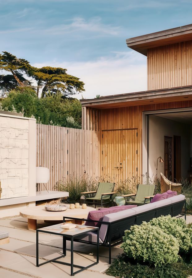 A refined contemporary outdoor courtyard or terrace adjoining a timber-clad house. The space features a built-in pale stone fireplace, outdoor lounge seating with black frames and muted pink cushions, a low round coffee table, side tables, ornamental grasses, and layered planting. Vertical timber screening, warm timber wall cladding, and broad roof overhangs create a sheltered, resort-like setting.