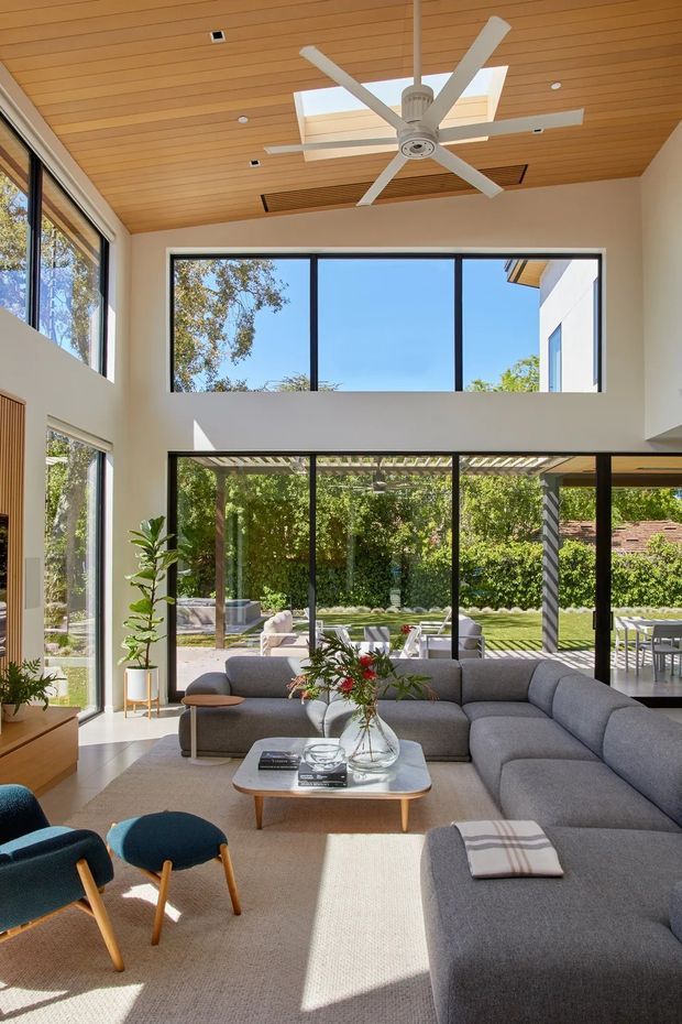 Bright contemporary living room with a double-height volume, expansive black-framed glazing, and a timber-lined ceiling fitted with a large ceiling fan and skylight. A large grey sectional sofa, textured neutral rug, wood coffee table, and teal accent chairs create a relaxed seating area. The glazing opens to a leafy landscaped outdoor area and covered patio.