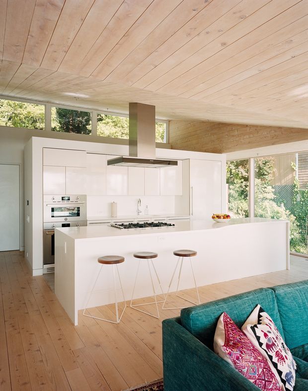 Bright open-plan kitchen and living area with a large white island, integrated white cabinetry, built-in stainless-steel appliances, gas cooktop, and a slim rangehood. Three stools line the island, while the foreground includes part of a teal sofa with patterned cushions. Extensive glazing brings in garden views, and the room is defined by warm light timber flooring and a matching timber-lined ceiling with a low, contemporary ceiling plane.