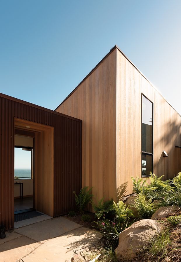 Exterior view of a contemporary timber-clad building with a sharp gabled roof and tall, narrow window. A dark corrugated-clad entry volume frames an open doorway, while native-looking planting and large rocks line the concrete path. A distant ocean horizon is visible through the entry, suggesting a coastal setting.