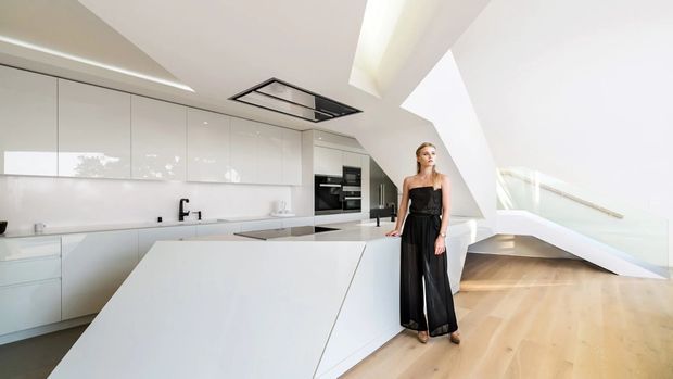 A bright, spacious contemporary kitchen with glossy white full-height cabinetry, a large angular white island, integrated black appliances, and a black undermount cooktop. Pale timber flooring extends toward a sculptural stairway and landing with glass balustrades. A large skylight and clerestory-style glazing bring in abundant natural light. A person stands beside the island.