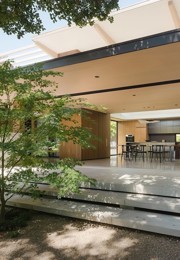 A modern open-plan kitchen and dining area opening onto a landscaped courtyard or garden. The space features broad white concrete or stone terraces and steps, warm vertical timber wall cladding and cabinetry, dark metal fascia and trim, large glazed openings, and a flat roof with a partially visible pergola or skylight structure. A dining island with stools and integrated kitchen appliances is visible beneath a minimalist linear pendant light. Mature leafy planting partially screens the architecture and casts dappled shadows across the terrace.