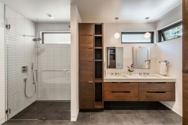 A contemporary bathroom featuring a walk-in tiled shower with frameless glass, a wall-mounted timber vanity with integrated basin, mirrored medicine cabinets, open shelving, and pendant lighting. The space has white horizontal wall tiles, dark grey floor tiles, warm wood cabinetry, and high horizontal windows.