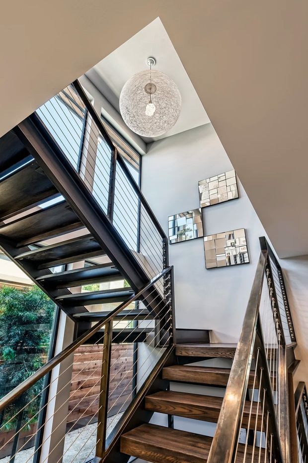 Interior view looking up a modern stairway with open-riser dark timber treads, dark metal stringers and cable railings. The stairwell is bright and airy, with tall glazing beside the stairs, a high angled ceiling, a large spherical pendant light, and decorative mirrored wall panels. Lush greenery is visible outside through the windows.