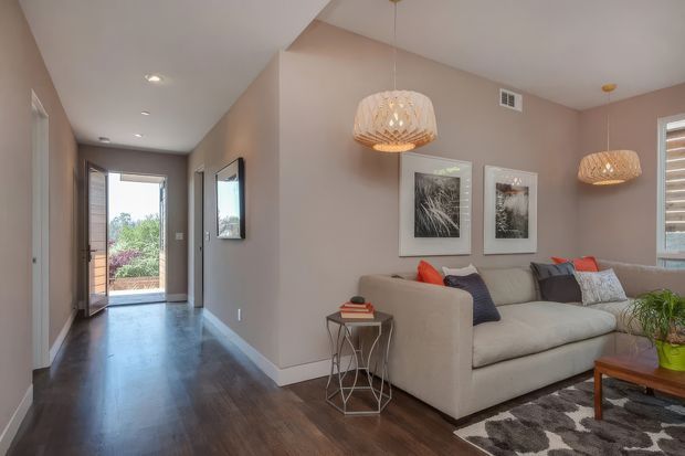 A warm contemporary living room viewed from an adjoining hallway, featuring dark timber flooring, taupe-painted walls, a large light neutral sectional sofa, patterned dark rug, framed artwork, woven pendant lights, and a timber coffee table. The hallway leads to an open exterior doorway, while a window with timber shutters provides additional daylight.