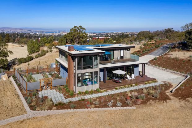 Aerial view of a contemporary multi-level house on a dry, landscaped sloping site. The home features broad glazing, dark metal framing, warm timber-toned vertical cladding, flat roof planes, terraces, and extensive paved outdoor areas. A distant urban and coastal-looking landscape is visible beyond the property.