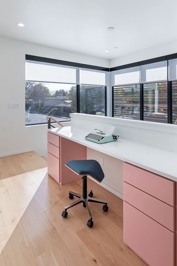 A bright, modern home office with a long white built-in desk wrapping beneath large corner windows. The desk incorporates extensive soft pink cabinetry and drawers, paired with a dark blue ergonomic stool and a mint-green typewriter. White roller blinds, black window frames, and timber flooring create a clean, contemporary workspace with leafy suburban views.