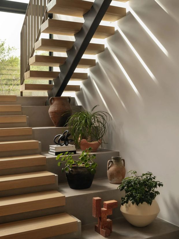 Interior view of a sculptural stairway with floating warm timber treads supported by a dark central steel beam. Concrete-grey stepped platforms, a pale plaster wall, vertical timber screening, and strong linear sunlight create a gallery-like setting with potted plants and ceramic vessels.