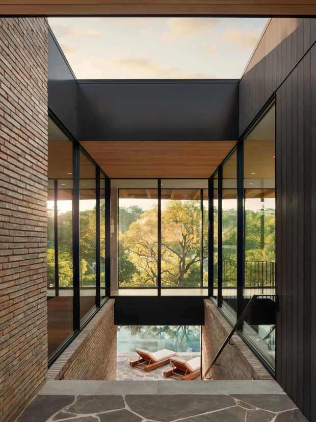 A dramatic interior architectural view looking down a glazed stairwell or landing toward a lower-level lounge and outdoor area. Exposed brick walls frame the space, with black metal-framed floor-to-ceiling glazing, dark vertical wall cladding, warm timber soffits, and a large overhead skylight or open roof section. A central mature tree is visible beyond the glazing, while the lower level contains lounge chairs on a stone-paved surface. The composition emphasizes strong vertical lines, transparency, layered levels, and natural light.