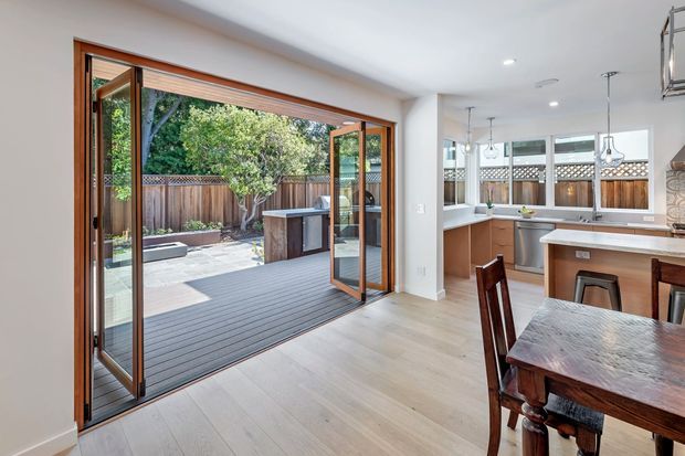 Bright open-plan kitchen and dining area with pale timber flooring, white walls, timber-framed bifold doors opening onto a timber deck, and a landscaped courtyard beyond. The kitchen features light cabinetry, a central island with pendant lights, and a timber dining table in the foreground.