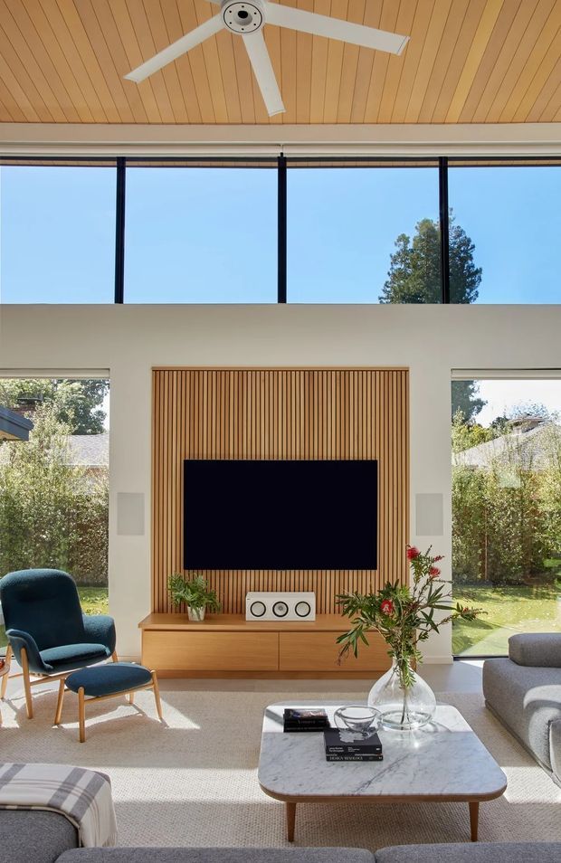 Bright contemporary living room with a double-height glazed wall, white structural walls, a warm timber-lined ceiling, and expansive windows overlooking greenery. A timber slatted feature wall surrounds a wall-mounted television above a built-in timber media console. The room includes a modern lounge, blue accent armchair and ottoman, marble-topped coffee table, indoor plants, and a large white ceiling fan.