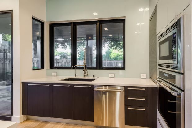 A contemporary galley-style kitchen with dark timber-toned cabinetry, a pale engineered-looking benchtop, integrated stainless-steel dishwasher, and built-in stainless-steel microwave and oven. A double-bowl sink sits beneath black-framed windows, with light walls and timber flooring creating a bright, practical interior.