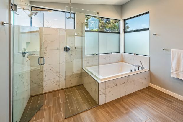Bright contemporary bathroom with a large frameless glass walk-in shower, wall-mounted shower fixtures, a freestanding-style bathtub enclosed in matching marble-look wall tiles, and broad corner windows. The room has warm wood-look tiled flooring, pale neutral walls, and abundant natural light.
