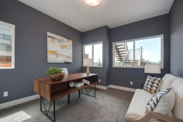 A compact, stylish living room or lounge with charcoal-grey walls, white trim, grey carpet, and a mid-century-inspired timber console on black metal legs. A white upholstered armchair with patterned cushions sits beside a floor lamp, while large windows provide abundant natural light and views of an exterior stair structure. Abstract artwork and restrained furnishings create a contemporary, refined atmosphere.