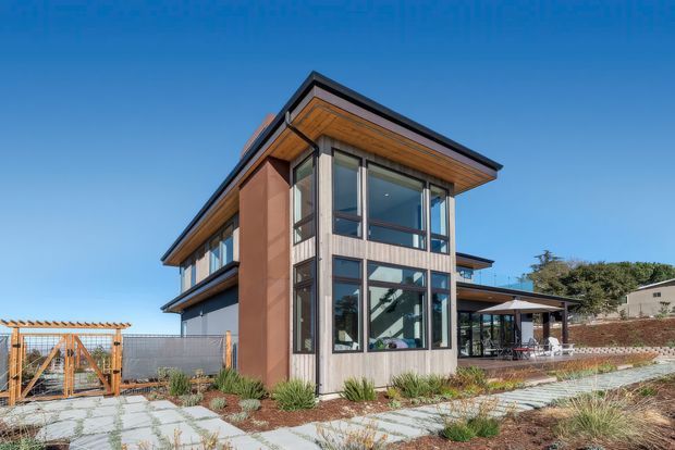 Exterior photograph of a contemporary multi-level residence with a prominent glazed corner volume, warm timber soffits, dark roof trim, and mixed vertical cladding. The house sits within a landscaped paved courtyard or garden, with an outdoor dining area and pergola visible to the side.
